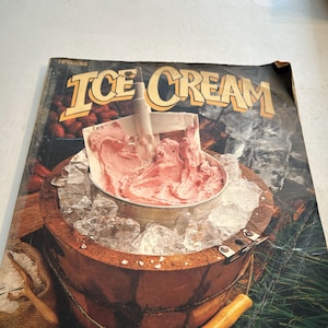 Puede incluir: Un libro de cocina vintage titulado "ICE CREAM" con una heladera de madera en uso. La heladera está llena de helado rosa y rodeada de hielo. También se ven un cubo de madera y una cuchara.