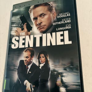 May include: A DVD case for the film The Sentinel, featuring Michael Douglas, Kiefer Sutherland, and Eva Longoria. The cover art displays the actors in suits, with two holding handguns, and the title text The Sentinel in large white letters.