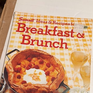 May include: A cookbook titled Sunset Ideas & Recipes for Breakfast & Brunch with a yellow and white checkered background. The cover features a large baked pancake topped with raspberries and mandarin oranges, alongside two glasses of orange juice.