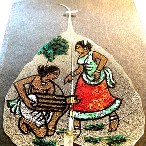 May include: A hand-painted leaf art piece depicting two figures in traditional clothing. The figures are painted in vibrant colors, including red, green, and black, on a translucent leaf. The leaf is mounted on a gray background.