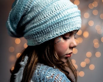 Könnte beinhalten: Eine junge Person trägt eine hellblaue Strickmütze mit einem lockeren Sitz und einen hellblauen Häkel-Schal. Die Mütze hat ein strukturiertes Strickmuster und der Schal hat ein zartes Lochmuster. Die Person hat langes braunes Haar und schaut nach unten.