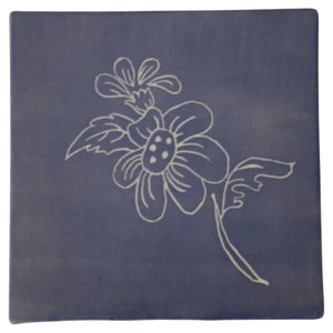 May include: A blue ceramic tile with a white floral design. The flower has five petals and a center with three dots. The tile has a slightly textured surface.