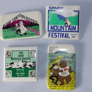 May include: Four vintage Sandy Mountain Festival buttons. The buttons are white with green, red, and blue accents. The buttons feature a bear, a mountain, and the text "Sandy Mountain Festival".