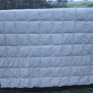 May include: A light gray, quilted blanket with a grid pattern, hanging over a wooden fence. The blanket is made of a soft, possibly down-filled material, and is evenly stitched to create square pockets. The background shows a natural outdoor setting.