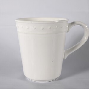 May include: A white ceramic mug with a raised design around the rim. The mug has a handle and a rounded bottom.