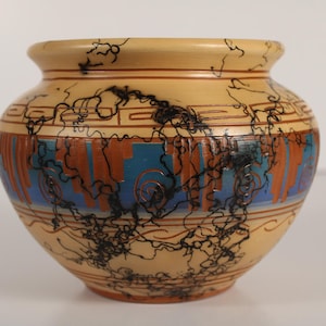 Puede incluir: Un jarrón de cerámica con una base de color beige, adornado con una banda de diseños abstractos azules, naranjas y negros. El jarrón tiene un cuerpo redondeado y un borde ligeramente ensanchado. El exterior presenta intrincado arte lineal negro.