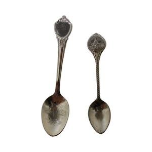 May include: Two silver souvenir spoons. The larger spoon has a decorative handle and a design on the bowl that says "St. Augustine, Florida". The smaller spoon has a round handle with a design of a building.