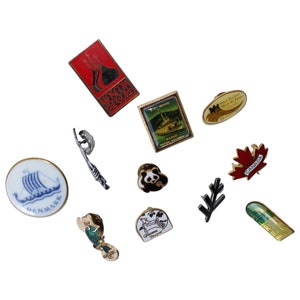 May include: A collection of ten enamel pins featuring various designs, including a panda, a maple leaf with the word "CANADA", a boat with the word "DENMARK", a fish, a building with the text "NAKLO", a building with the text "MOSCOW TREASURES", a building with the text "WHISTLER MOUNTAIN", a building with the text "BANFF JASPER", and a building with the text "EL ESTOR COLOMBIA".