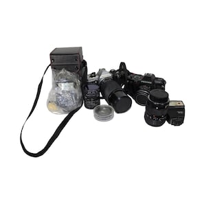 Puede incluir: Una bolsa de cámara negra con una correa, una cámara Pentax negra y plateada con un objetivo, un objetivo negro y un flash negro.