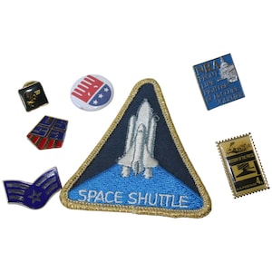 May include: A collection of seven patches and pins, including a blue and white space shuttle patch, a red, white, and blue button with stars, a blue and gold pin with a star, a blue and gold pin with a building, a blue and gold pin with a gun, a red, white, and blue pin with a symbol, and a gold stamp with a picture of a hand holding a pen.
