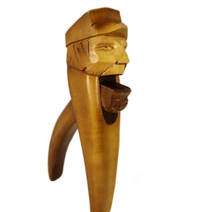 Puede incluir: Un cascanueces de madera tallado en forma de cabeza de persona con sombrero. El cascanueces es marrón y tiene una pequeña abertura para colocar las nueces.