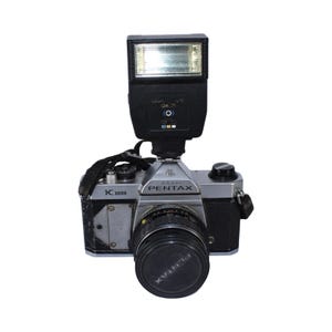 May include: A vintage silver and black Pentax K1000 35mm film camera with a flash attachment. The camera has a black lens cap and a black strap. The flash is black with a clear front.