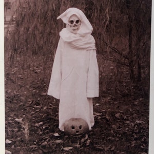 Puede incluir: Un niño disfrazado de fantasma para Halloween, con una túnica blanca con capucha y una máscara de calavera. El niño está de pie en un área boscosa con una calabaza tallada a sus pies.