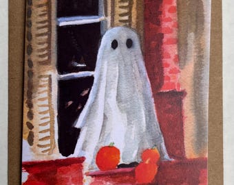 Può includere: Un'illustrazione ad acquerello per biglietto d'auguri raffigurante un fantasma bianco in piedi su un portico di mattoni rossi davanti a una finestra scura con persiane color sabbia. Tre piccole zucche arancioni sono alla base del fantasma.