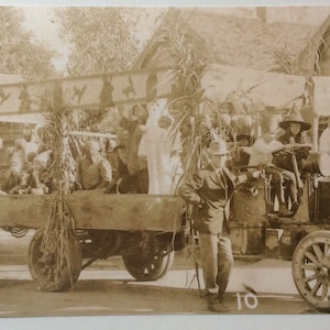 May include: Sepia-toned photograph of a vintage parade float, likely from the early 20th century. The float is a large truck bed decorated with foliage and carrying several people. A smaller car is in front of the float. The number "10" is visible.
