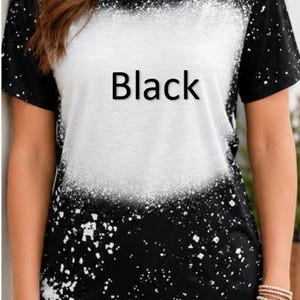 May include: A short-sleeved t-shirt featuring a black base color with a large, central white bleached-effect area and scattered white paint-like splatters throughout. The word Black is printed in black text across the center of the shirt.