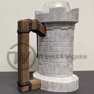 Puede incluir: Una taza de cer&aacute;mica blanca con forma de castillo con un asa de madera marr&oacute;n. La taza tiene una textura de piedra y una bandera azul y blanca colgando del asa.