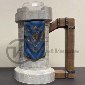 Puede incluir: Una taza de cer&aacute;mica blanca con forma de torre de piedra con una pancarta azul y dorada. La taza tiene un asa de madera con detalles met&aacute;licos.