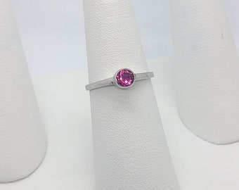 Op de afbeelding: Een zilveren ring met een roze edelsteen op een witte ringstandaard.