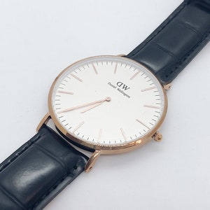 Könnte beinhalten: Eine roségoldene Uhr mit weißem Zifferblatt und schwarzem Lederarmband. Das Zifferblatt der Uhr hat schwarze römische Ziffern als Stundenmarkierungen und den Text "DW Daniel Wellington" aufgedruckt.
