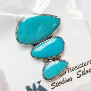 May include: A turquoise pendant with three stones set in sterling silver. The pendant is in a clear plastic bag with a white card that says "Tarnish Resistant Sterling Silver" and has a logo of a stylized "MK".