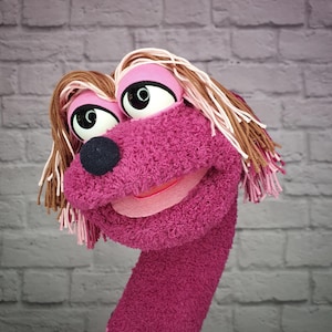 Könnte beinhalten: Eine magentafarbene Handpuppe mit texturierter, flauschiger Oberfläche, großen weißen Augen mit schwarzen Pupillen, einer runden schwarzen Nase und einem breiten Mund. Die Puppe hat langes, geschichtetes braunes und hellrosa Wollhaar.