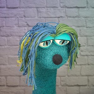 Könnte beinhalten: Eine blaugrüne Handpuppe mit texturierter, flauschiger Oberfläche, einem runden schwarzen Nasenknopf und großen, herabhängenden Augenlidern über katzenartigen Augen mit blauen Iris. Die Puppe hat langes, geschichtetes Garnhaar in Blau, Grün und Gelb.