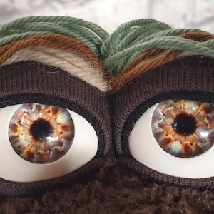 Könnte beinhalten: Ein Plüschtier mit zwei großen, realistischen Glasaugen mit braunen und orangefarbenen Iris. Die Augen sind von dunkelbraunen Stofflidern umrahmt und mit dickem, geschichtetem Garnhaar in Salbeigrün, Creme und Dunkelbraun bedeckt.