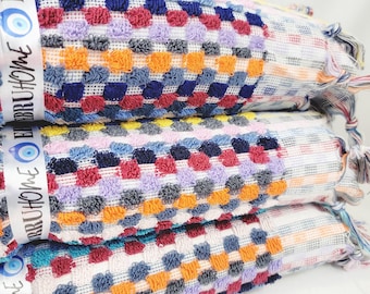 May include: Three colorful, textured towels stacked on top of each other. The towels have a checkered pattern in shades of blue, orange, purple, red, and gray. Each towel is wrapped with a white ribbon that has the text "Ebru Home" and an eye symbol.