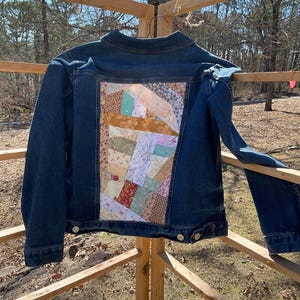 May include: A blue denim jacket with a patchwork design on the back. The patchwork is made of various fabrics in shades of brown, pink, green, and white.