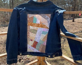 May include: A blue denim jacket with a patchwork design on the back. The patchwork is made of various fabrics in shades of brown, pink, green, and white.