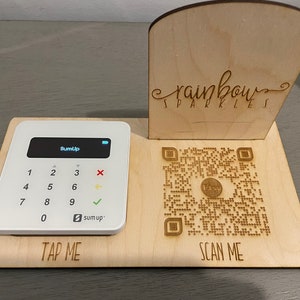 May include: A wooden stand with a white card reader and a QR code. The stand has the text "TAP ME" and "SCAN ME" engraved on it. The card reader has the text "SumUp" on the screen. The QR code has the text "Etsy" in the centre.