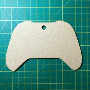 May include: A brown cardboard cutout of a video game controller with a hole in the top for hanging. The cutout is on a green cutting mat with white grid lines.