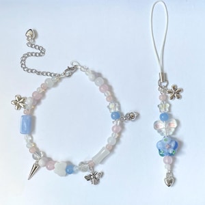 May include: A set of two delicate beaded bracelets with a pastel color scheme. One bracelet is a charm bracelet with a bee, flower, and cloud charm. The other bracelet is a phone charm with a flower and butterfly charm.
