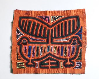 May include: A colorful textile panel with a geometric design of two stylized birds with outstretched wings. The panel is made of orange fabric with black, green, blue, pink, and yellow embroidery.