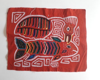 May include: A red fabric wall hanging with a colorful embroidered design of two fish. The fish are outlined in white and filled with various colors, including black, red, yellow, blue, and green.