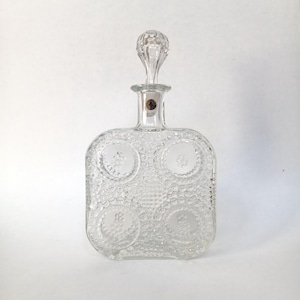 Puede incluir: Decantador de vidrio transparente con cuerpo cuadrado y tapón decorativo. El decantador presenta una superficie texturizada con cuatro diseños circulares. El tapón tiene forma de lágrima. Una pequeña etiqueta es visible en el cuello del decantador.