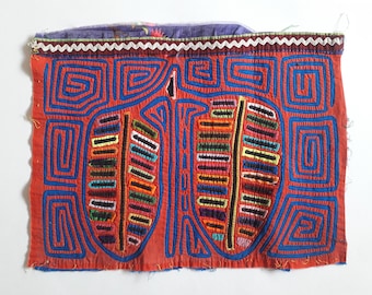 May include: A vibrant textile piece featuring a red background with blue geometric patterns and two central leaf-like shapes. The leaves are filled with colorful horizontal stripes. The top edge has a purple floral fabric and white trim.
