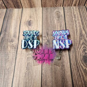 Könnte beinhalten: Zwei glitzernde blaue und violette Badge-Rollen mit dem Text "Sounds like a DSP" und "Sounds like a NSP" darauf. Die Badge-Rollen sind an klaren Acryl-Anhängern mit einem Stethoskop und einem Herzschlag-Motiv sowie dem Text "Crafts by a Nurse" befestigt.