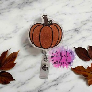 Könnte beinhalten: Ein transparenter Glitzer-Badgehalter mit einem funkelnden orangefarbenen Kürbisdesign. Der Badgehalter hat eine silberne einziehbare Rolle und einen silbernen Clip. Der Text "Crafts by a Nurse" ist im Hintergrund sichtbar.