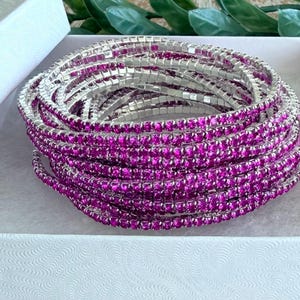 May include: A stack of multiple stretch bracelets with silver-colored metal and vibrant pink rhinestones. The bracelets are coiled inside a white box with a textured pattern, creating a sparkling and eye-catching display.