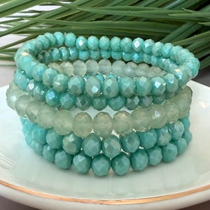 May include: Stack of four beaded bracelets in shades of turquoise and pale green. The bracelets are made of faceted beads, creating a sparkling effect. The bracelets are displayed on a white plate with a gold rim, with green foliage in the background.