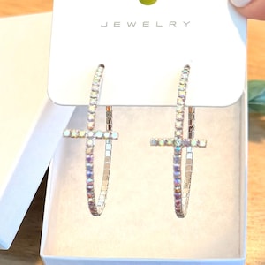 May include: Silver-toned hoop earrings with cross pendants, presented in a white box. The earrings are adorned with iridescent rhinestones. The word "JEWELRY" is printed on the white card.