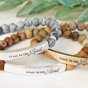 May include: Three beaded bracelets with silver and gold metal charms that say "Trust in the Lord". The bracelets are made with gray, brown, and beige stones.