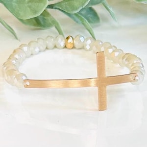 May include: A gold cross charm bracelet with white iridescent beads. The cross is a simple, minimalist design.