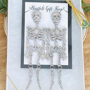 May include: A pair of silver-toned skeleton earrings featuring iridescent rhinestones throughout the body and head. The earrings are displayed on a white card with the text Hartfelt Gift Shop, inside a white gift box with a gold ribbon.