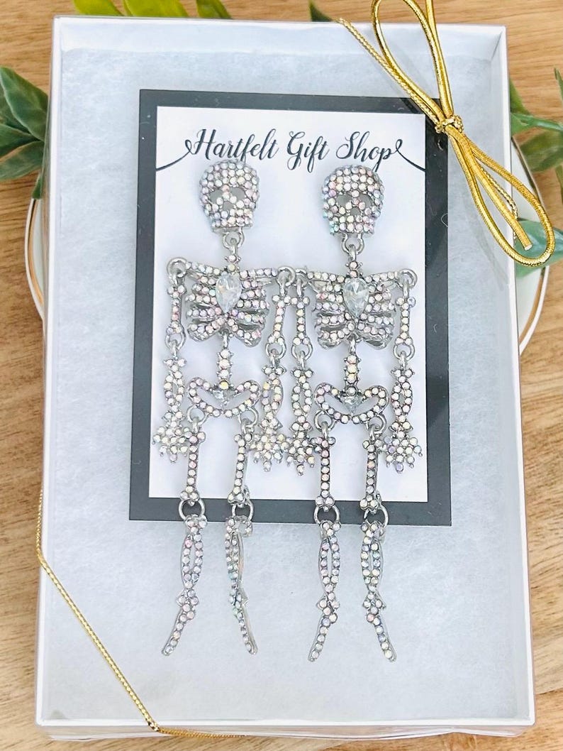 May include: A pair of silver-toned skeleton earrings featuring iridescent rhinestones throughout the body and head. The earrings are displayed on a white card with the text Hartfelt Gift Shop, inside a white gift box with a gold ribbon.