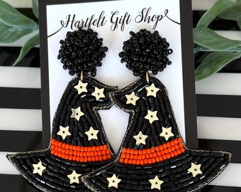 May include: A pair of beaded earrings shaped like witch hats on a Hartfelt Gift Shop display card. The hats are black with orange bands and gold star accents. Each earring features a round black beaded stud at the top and a wide brim.