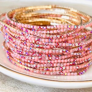 Może przedstawiać: Stos różowych i złotych bransoletek z kryształkami. Bransoletki wykonane są z małych, kwadratowych kryształków i mają delikatną, złotą podstawę z drutu.