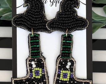 May include: A pair of beaded earrings featuring a black witch hat top connected to a black boot bottom. The boots have green and black striped shafts, a purple and lime green square buckle detail, and white accents, all outlined in metallic gold thread.